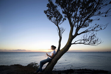 Junge Frau lehnt an einem Baum in der Abenddämmerung - ISF18134