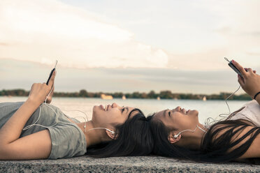 Young women lying head to head at waterfront listening to music through earphones - ISF18468