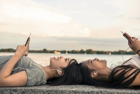 Junge Frauen liegen Kopf an Kopf am Wasser und hören Musik über Kopfhörer - ISF18468