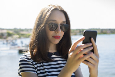 Italy, Lake Garda, portrait of young woman wearing sunglasses using smartphone - GIOF04048