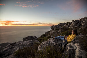 South Africa, Cape Town, Table Mountain, woman sitting on a rock drinking wine at sunset - DAWF00686