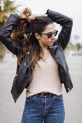 Spain, Barcelona, young woman wearing leather jacket doing her hair - MAUF01569
