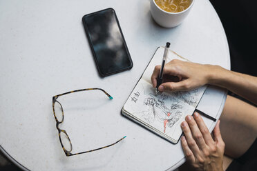 Hands of woman sketching in notebook, glasses, tea and smartphone on table - KKAF01263