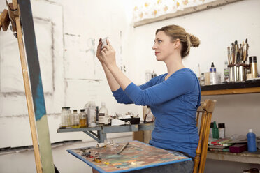 Mid adult woman photographing oil painting in artist's studio - ISF18795