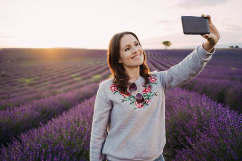 Frankreich, Valensole, Porträt einer lächelnden Frau, die ein Selfie vor einem Lavendelfeld bei Sonnenuntergang macht - GEMF02221