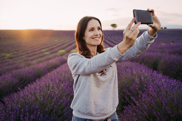 Frankreich, Valensole, Porträt einer lächelnden Frau, die ein Selfie vor einem Lavendelfeld bei Sonnenuntergang macht - GEMF02222