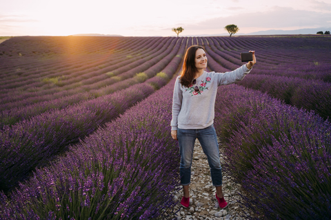 Frankreich, Valensole, lächelnde Frau macht Selfie auf Lavendelfeld bei Sonnenuntergang, lizenzfreies Stockfoto