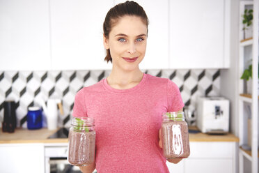 Portrait of smiling young woman with two glasses of raspberry smoothie in the kitchen - ABIF00795