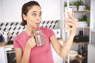 Portrait of young woman taking selfie with smartphone in the kitchen while drinking smoothie - ABIF00796