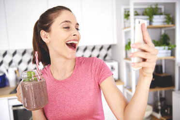 Portrait of laughing young woman with glass of smoothie taking selfie with smartphone in the kitchen - ABIF00797