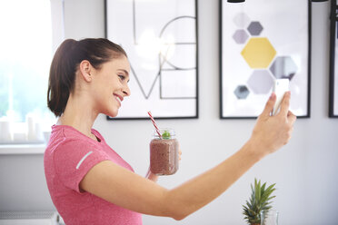 Smiling young woman with smoothie taking selfie with smartphone in the kitchen - ABIF00799