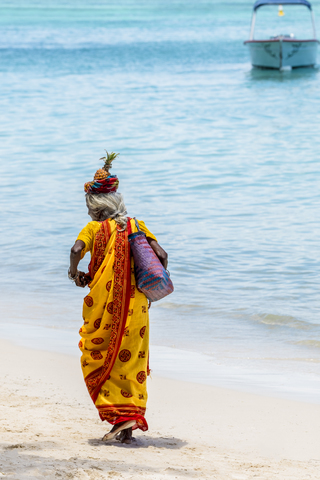 Mauritius, Trau-aux-Biches, einheimische Frau im Sari und mit Ananas auf dem Kopf beim Spaziergang am Strand, lizenzfreies Stockfoto