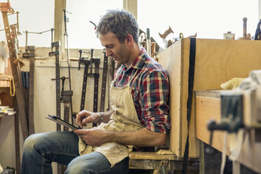 Ein Restaurator für antike Möbel sitzt in seiner Werkstatt und benutzt ein digitales Tablet. - MINF03051