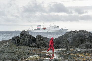 Mann, der am Meer in der Antarktis an Felsen entlang geht, im Hintergrund die Akademik Sergey Vavilov, ein russisches Polarforschungsschiff. - MINF03065