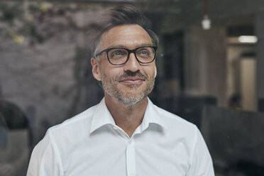 Porträt eines lächelnden Mannes mit Bartstoppeln hinter einer Fensterscheibe, der ein weißes Hemd und eine Brille trägt - PNEF00823