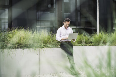 Smiling businessman using laptop outside office building - UUF14683