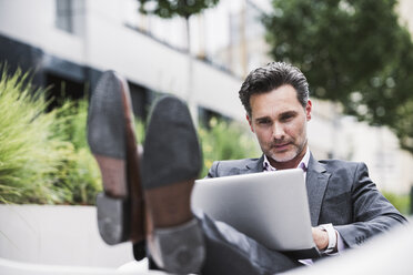 Relaxed businessman using laptop outside office building - UUF14691