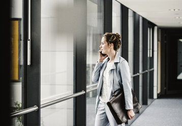 Businesswoman on cell phone looking out of window in office passageway - UUF14699