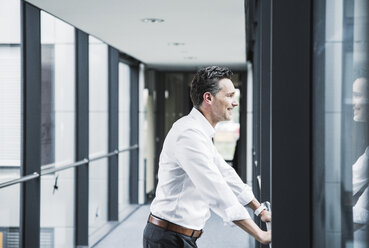 Smiling businessman looking out of window in office passageway - UUF14723