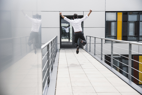 Ein jubelnder Geschäftsmann springt auf dem Skywalk eines Bürogebäudes, lizenzfreies Stockfoto