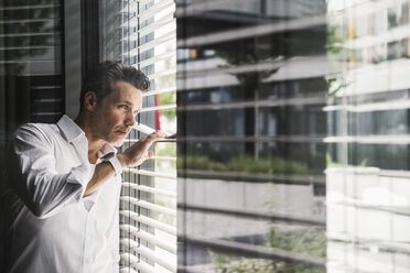 Geschäftsmann späht durch die Jalousien am Fenster im Büro - UUF14737