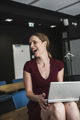 Laughing businesswoman in office wearing burgundy dress using laptop - UUF14741