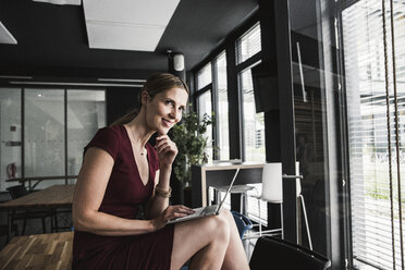 Smiling businesswoman in office wearing burgundy dress using laptop - UUF14743