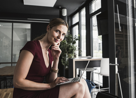 Geschäftsfrau im Büro in burgunderrotem Kleid mit Laptop, lizenzfreies Stockfoto