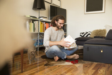 Junger Mann sitzt auf dem Boden im Wohnzimmer und packt ein Geschenk aus - MFRF01161