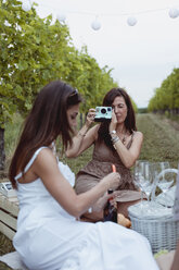Friends having picnic in a vineyard, one woman taking pictures with instant camera - MAUF01625