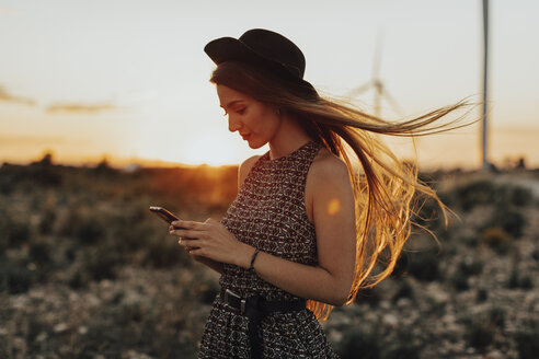 Porträt einer jungen Frau, die bei Sonnenuntergang auf ihr Mobiltelefon schaut - OCAF00338