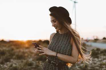 Porträt einer jungen Frau, die bei Sonnenuntergang auf ihr Mobiltelefon schaut - OCAF00339