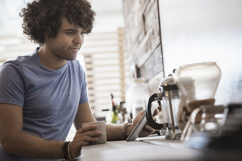 Ein junger Mann sitzt mit einer Tasse Kaffee in der Hand vor einem digitalen Tablet. - MINF03143