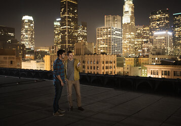 Zwei Männer auf einem Dach mit Blick auf Los Angeles bei Nacht, die auf ein Smartphone schauen. - MINF03148