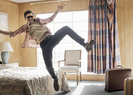 Ein junger Mann mit Sonnenbrille springt in einem Motelzimmer in die Luft. - MINF03153