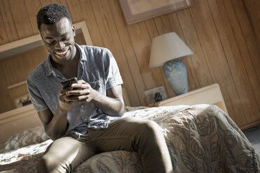 Ein junger Mann in einem Motelzimmer überprüft sein Smartphone. - MINF03162