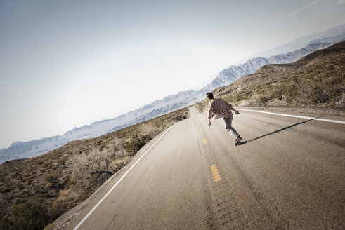 Ein junger Mann fährt auf einem Skateboard eine geteerte Straße in der Wüste hinunter. - MINF03175
