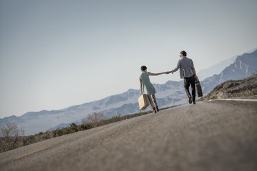 A young couple, man and woman walking hand in hand on a tarmac road in the desert carrying cases. - MINF03177