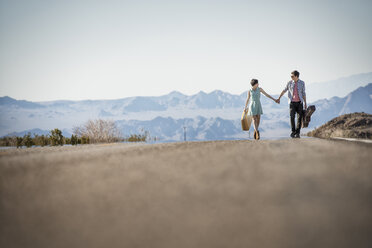 A young couple, man and woman walking hand in hand on a tarmac road in the desert carrying cases. - MINF03178