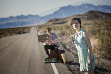 Ein junges Paar, Mann und Frau, trampen auf einer asphaltierten Straße in der Wüste, mit einem Schild, auf dem 