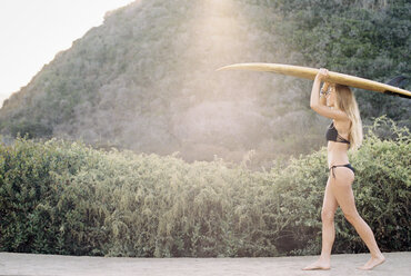 A woman in a black bikini walking down a path carrying a surfboard on her head. - MINF03222