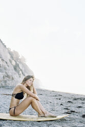 Eine Frau im Bikini sitzt auf ihrem Surfbrett an einem Sandstrand. - MINF03224