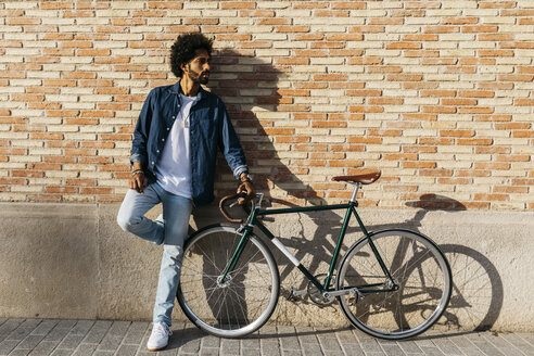 Young man with racing cycle leaning against brick wall - JRFF01746