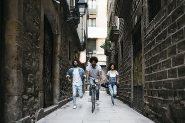 Spain, Barcelona, man riding bicycle down an alley while his friends running behind him - JRFF01768