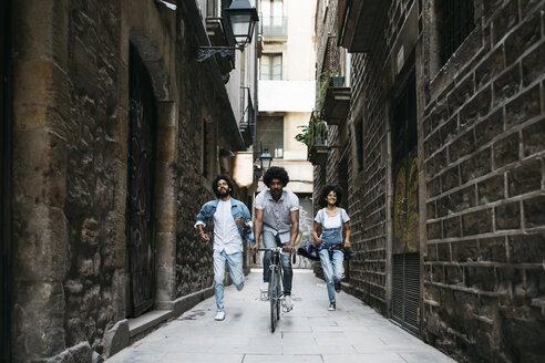 Spain, Barcelona, man riding bicycle down an alley while his friends running behind him - JRFF01768