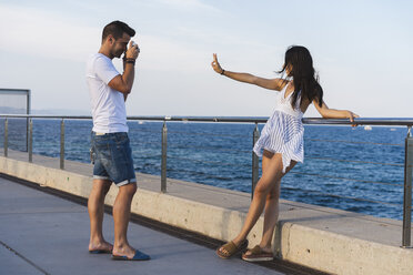 Young urban couple taking pictures by the sea, woman making peace sign - AFVF01168