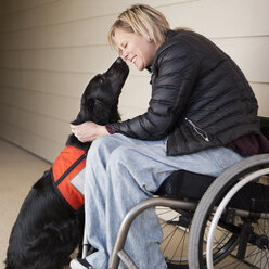 A mature woman wheelchair user with her service dog, a black Labrador, leaning in towards each other. - MINF03317