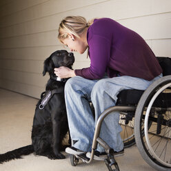 A mature woman wheelchair user stroking her black Labrador service dog and making eye contact with the dog. - MINF03318