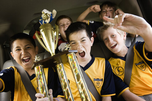 Eine Gruppe von Jungen in Fußballtrikots, die einen Pokal halten und einen Sieg feiern, in einem Mannschaftsbus. - MINF03390