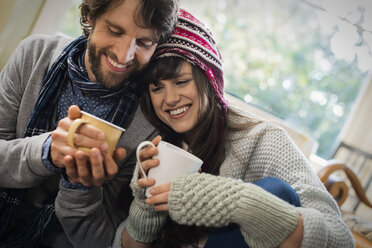 A couple, man and woman sitting close together warming up with their hands around mugs. - MINF03451